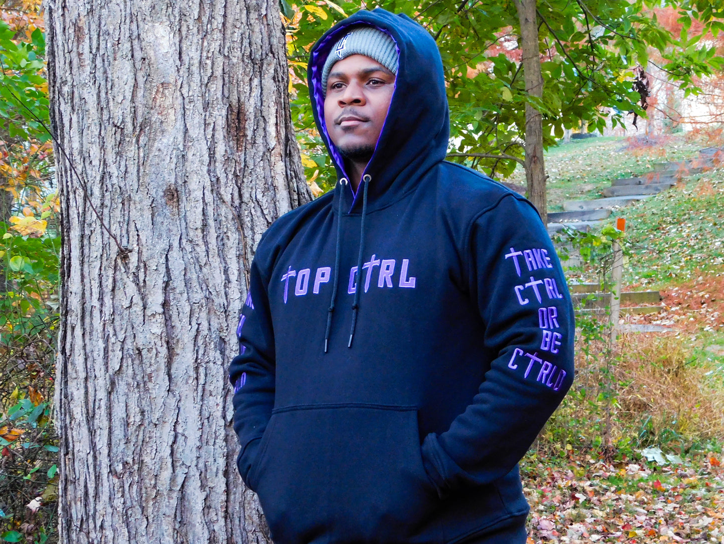 Person wearing a black and purple hoodie with text, standing next to a tree in a forest setting.