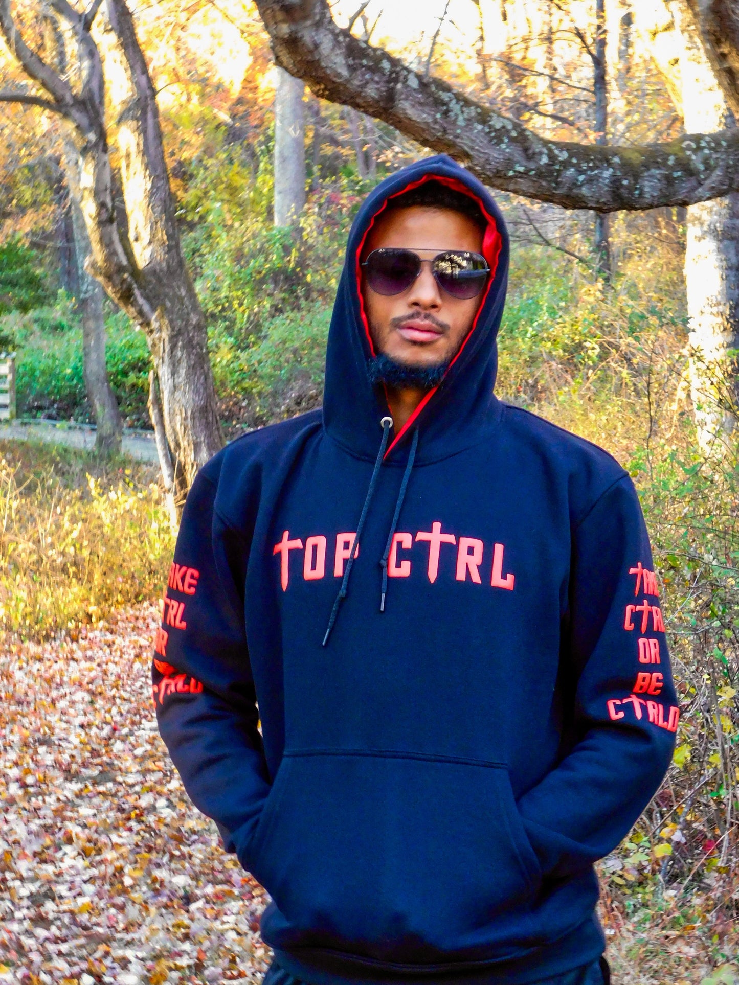 Person wearing a black and red hoodie with red text standing in a forest during autumn.